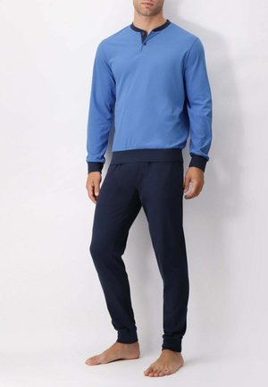 Uomo che indossa una maglia henley a maniche lunghe blu con colletto e polsini neri e pantaloni jogger neri, in piedi a piedi nudi su uno sfondo bianco.