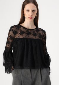 Blusa nera con un corpino in pizzo trasparente e testurizzato con motivi floreali. Le maniche lunghe presentano orli svasati, abbinata a pantaloni grigi a vita alta.