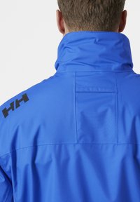 Heldere blauwe waterdichte jas met hoge kraag. Bevat een zwart logo op de schouder en een patchzak op de rug. Soepele textuur.