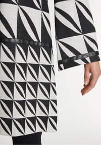 Černobílé šaty s vzorem ve tvaru trojúhelníků, hladkou texturou a černým lemem na okrajích a rukávech.