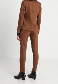 Brun skräddarsydd kavaj med guldknappar, matchad med passande skräddarsydda byxor. Svarta ankelskor fulländar looken. Slät tygtextur.