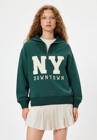Sudadera verde con un alto cuello, media cremallera y letras en crema "NY DOWNTOWN". Llevada sobre una falda plisada en crema. Tejido suave, diseño informal.