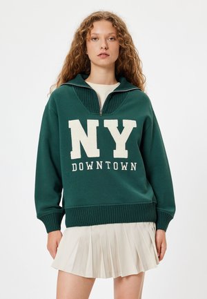 Groene sweatshirt met een hoge kraag, halve ritssluiting en crèmekleurige tekst "NY DOWNTOWN". Draagt over een crèmekleurige geplisseerde rok. Zachte stof, casual ontwerp.