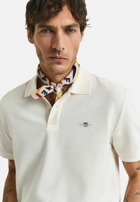 Weißes Poloshirt aus strukturiertem Stoff; hat ein kleines, gesticktes Logo. Ein gemusterter Schal in Rot, Blau und Gelb ist am Hals gebunden.