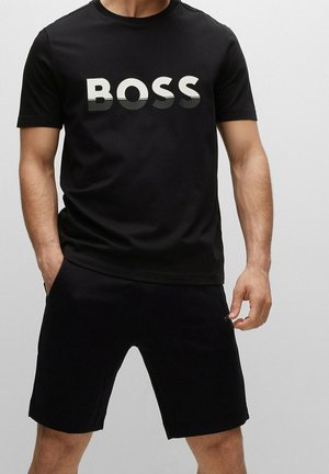Mann trägt schwarzes T-Shirt mit auffälligem weiß-grauem Farbverlauf und dem Schriftzug "BOSS", kombiniert mit schwarzen Shorts vor einfachem hellem Hintergrund.