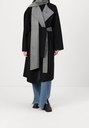 Zwart gesealde jas met grijze brede revers en sjaal, gecombineerd met blauwe jeans met wijde pijpen en zwarte loafers tegen een effen achtergrond.