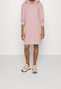 Vestido con capucha en suave tela rosa con un bolsillo frontal y cordón. Lleva unos zapatos deportivos gruesos en beige y blanco con detalles en naranja.