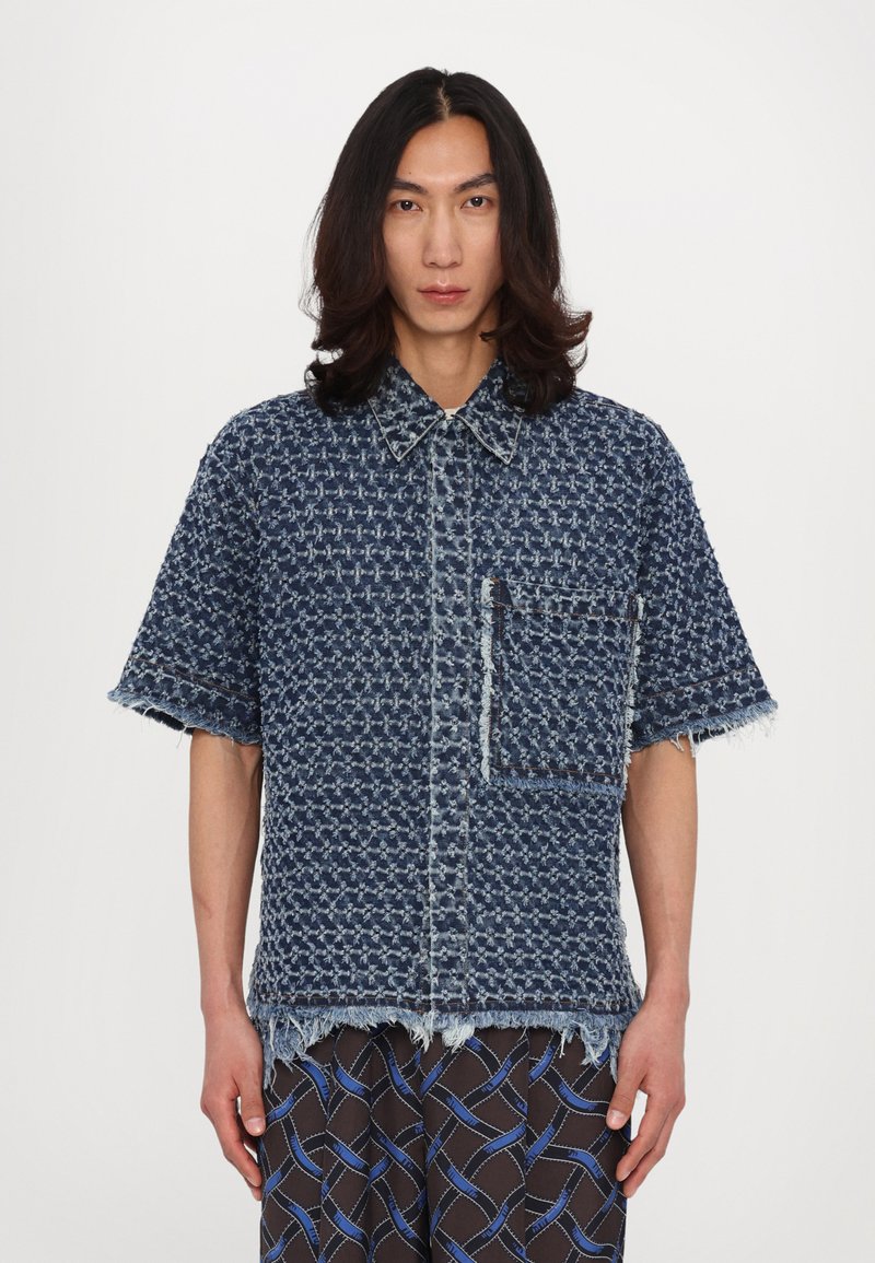 Denim shirt with a textured blue pattern, short sleeves, frayed hem, and a single chest pocket. Features contrasting stitching detail.