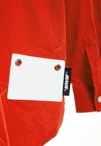 Camisa de algodón roja con una etiqueta blanca que cuenta con ojales, detalles de logotipo en relieve y una etiqueta de marca negra en la costura lateral.
