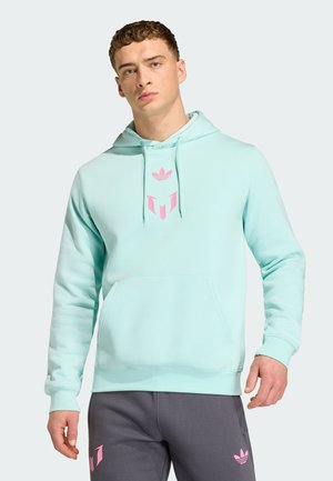 Joven con sudadera con capucha azul clara con logo rosa y pantalones gris oscuro con logo rosa a juego, de pie frente a un fondo liso claro.
