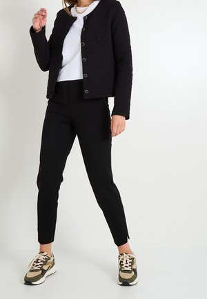 Femme portant une veste noire texturée boutonnée, une chemise blanche, un pantalon noir et des baskets vert olive, debout sur fond blanc.