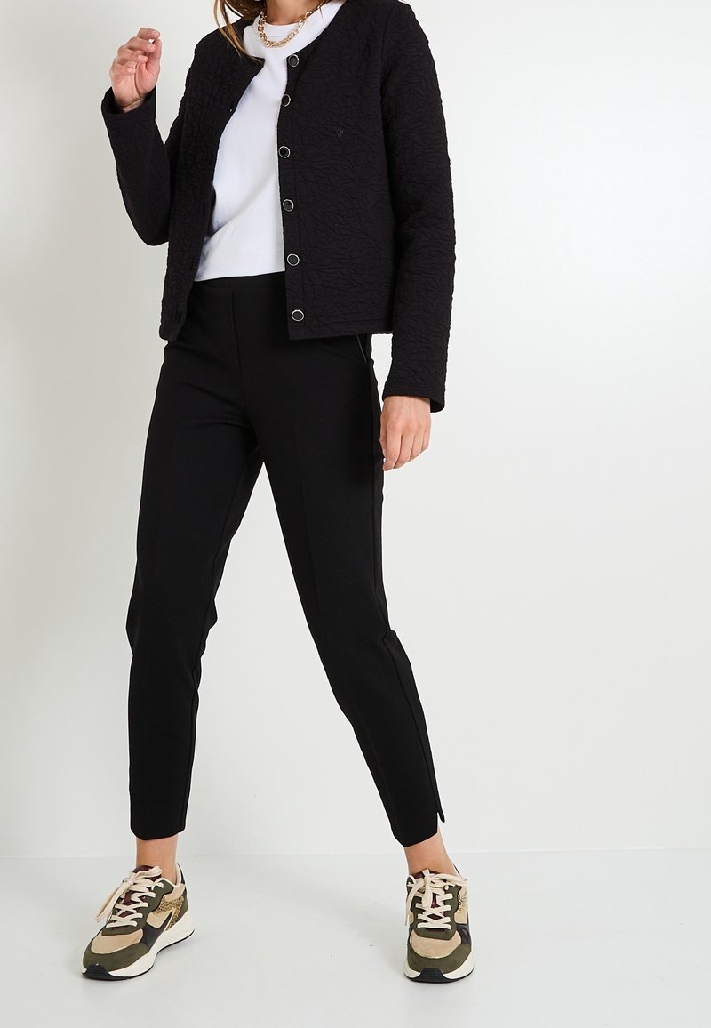 Femme portant une veste noire texturée boutonnée, une chemise blanche, un pantalon noir et des baskets vert olive, debout sur fond blanc.