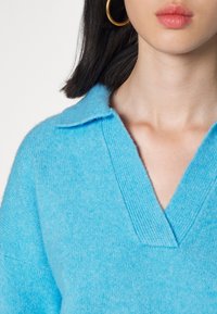 Hellblauer Pullover mit V-Ausschnitt und Kragen, der eine weiche Textur und gerippte Details entlang des Halsausschnitts zeigt.