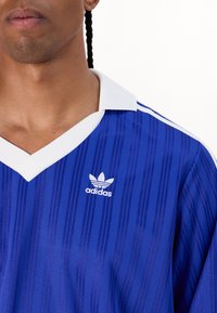 Vīrietis, ģērbts zilos Adidas sporta krekliņos ar baltu apkakli un izšūtu logotipu uz krūtīm, uz balta fona.