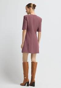 Mujer vestida con un vestido corto de manga corta en color malva, con costuras verticales y botas de cuero marrón de tacón hasta la rodilla, de pie frente a un fondo simple.