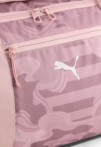 Κοντινή λήψη υφάσματος με ροζ καμουφλάζ μοτίβο, με ανοιχτό ροζ φερμουάρ και λευκό λογότυπο Puma σε μια τσέπη.