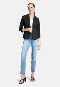 Zwarte blazer met een sjaalkraag, enkele knoopsluiting en driekwartmouwen, gedragen over een witte top en lichtblauwe jeans.