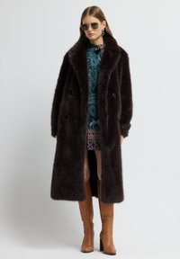 Abrigo de piel sintética marrón con un gran cuello y diseño de doble botonadura, combinado con un vestido estampado y botas de caña alta en color marrón claro.