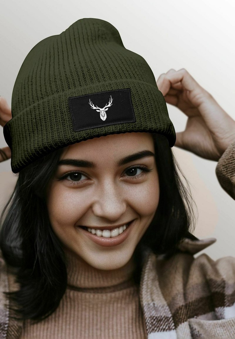 Olive green knitted beanie with a textured design and a black patch featuring a deer graphic. Soft material and fold-over cuff.