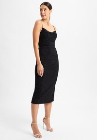 Schwarzes Midikleid mit dünnen Trägern, einem Cowl-Ausschnitt und schimmernden Details. Eng anliegende Form betont die Silhouette. Kombiniert mit silbernen Riemchenheels.