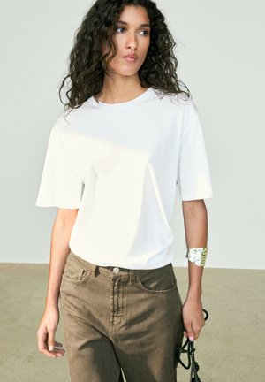 Donna con lunghi capelli ricci indossa una semplice maglietta bianca, jeans marroni, bracciale a polsino d’argento e tiene un cinturino nero, in piedi all’interno.