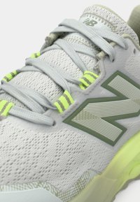 Hellgrauer Sportschuh mit einem strukturierten Mesh-Obermaterial, grünen Akzenten, reflektierenden Details und einer dezenten gemusterten Sohle. Ausgestattet mit hellgelben Schnürsenkeln.