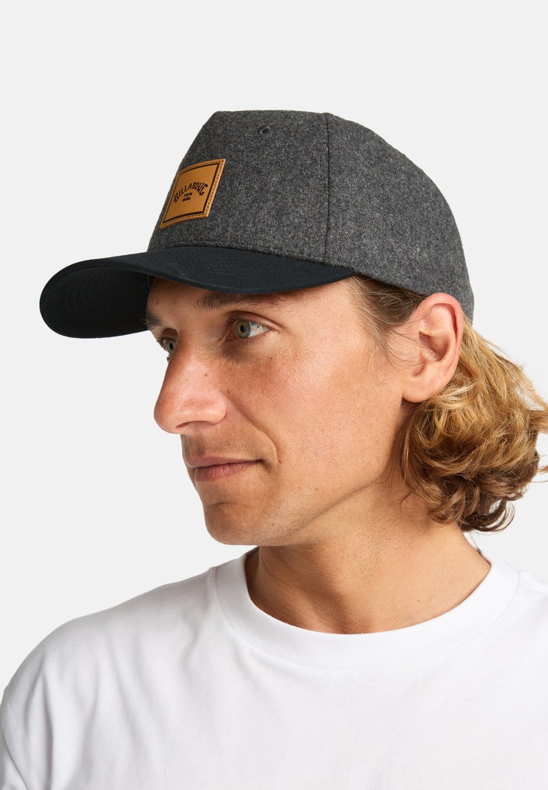 Gorra de lana gris con visera negra, que presenta un parche de cuero marrón en la parte frontal. Vista en primer plano de perfil.