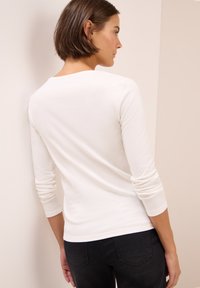 Femme aux cheveux bruns courts portant une chemise blanche à manches longues et un pantalon noir, vue de dos contre un mur de coin beige.