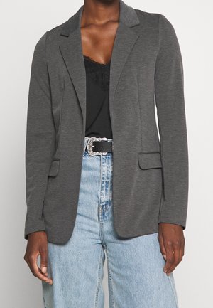 Personne portant un blazer gris foncé sur un haut noir orné de dentelle, associé à un jean taille haute bleu clair et une ceinture noire avec une boucle argentée.