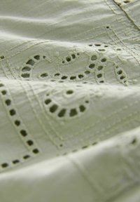 Jasnozielony materiał z haftowanymi wzorami paisley, z wyciętymi detalami i teksturowaną powierzchnią. Gładki i nieco pomarszczony wygląd.