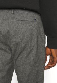 Gros plan sur une personne portant un pantalon en laine gris avec une poche arrière et un pull noir, main détendue sur le côté.