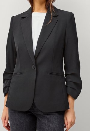 Femme portant un blazer noir cintré à un seul bouton sur un haut blanc et un jean foncé sur un fond clair uni.