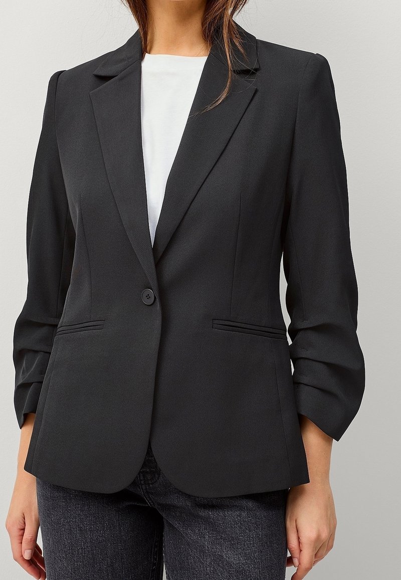 Femme portant un blazer noir cintré à un seul bouton sur un haut blanc et un jean foncé sur un fond clair uni.