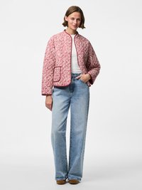 Giacca trapuntata con un motivo floreale in rosso e bianco, presenta un collo rotondo, maniche lunghe e una tasca frontale. Indossata sopra un top bianco e jeans.