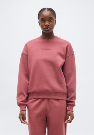 Kobieta z warkoczami ubrana w pasujący, stonowany różowy sweatshirt i spodnie, stojąca na tle prostego jasnoszarego tła.