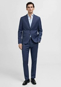 Traje azul marino con chaqueta de un solo botón, solapas en pico y pantalones a juego. Camisa azul clara por debajo, combinada con zapatos negros.
