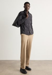 Camicia a righe nere con ricami bianchi, pantaloni beige, scarpe nere e una borsa tote nera su uno sfondo neutro.