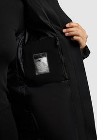 Jachetă neagră cu un material exterior neted, căptușeală matlasată la interior, buzunar cu fermoar și etichetă de întreținere vizibilă în interior pe partea dreaptă.