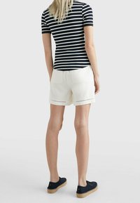 Kobieta stojąca tyłem, ubrana w czarno-białą paskowaną koszulę, białe szorty z tylnymi kieszeniami oraz czarne espadryle.