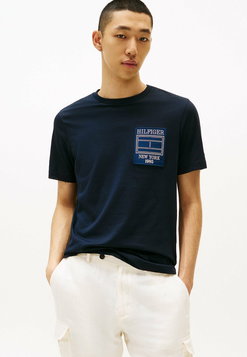 T-shirt in cotone blu navy con collo a giro, caratterizzato da un patch logo ricamato blu e bianco sul petto. Texture liscia, vestibilità casual.