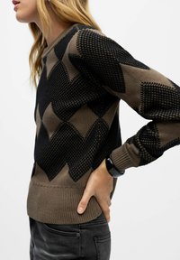 Strikket sweater i jordnære nuancer med et sort chevron-mønster, ribbede manchetter og kant, med et struktureret design, vist fra siden.