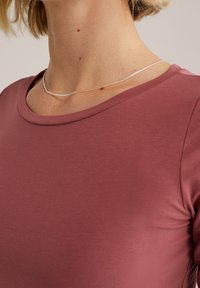 Top aderente color marrone scuro con scollatura rotonda e maniche corte, realizzato in un tessuto liscio. Una delicata collana in argento è visibile.