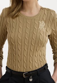 Beige svetr s pleteným vzorem a kulatým výstřihem, dekorativními knoflíky na rameni a malým logem vyšitým na hrudi.