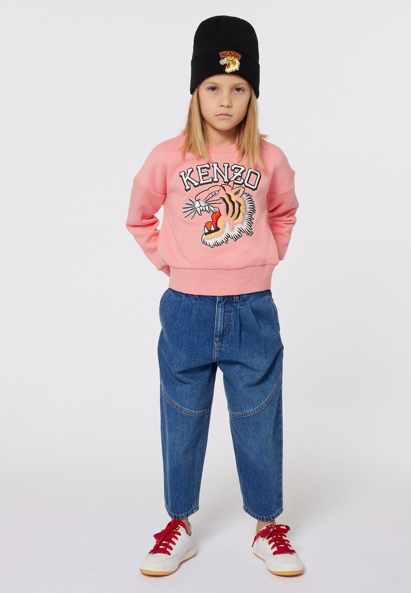 Sweatshirt rose avec le graphique "KENZO" et un tigre, pantalon en denim baggy bleu, bonnet en tricot noir, et baskets blanches avec des lacets rouges.