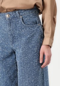 Sinine struktuurne denim pükste kõrge talje, millel on eesmine tasku, metallist nupuga kinnitamine ja valgete täppidega muster.