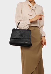 Borsa nera con texture, di forma rettangolare, dotata di una chiusura rotonda e di una tracolla regolabile. Design minimalista con accenti metallici.