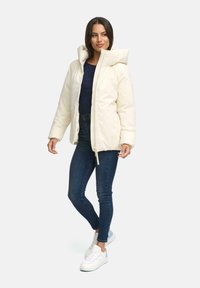 Vrouw in een crèmekleurige pufferjack, donkerblauwe jeans, marineblauw shirt en witte sneakers, staande tegen een effen lichte achtergrond.