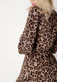 Brązowa sukienka w panterkę z długimi, bufiastymi rękawami, talią podkreśloną paskiem i zwiewną tkaniną, prezentująca odważny zwierzęcy wzór.