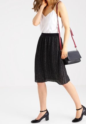 Jupe plissée noire avec une finition texturée, associée à un haut blanc à col en V et des chaussures à talons noires. Accessoires comprenant un sac à main noir avec une sangle bordeaux.