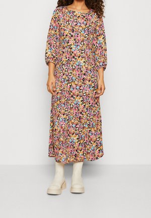 Femme portant une longue robe à motif floral avec des fleurs roses, bleues, jaunes et oranges, associée à des bottes épaisses couleur écrue.